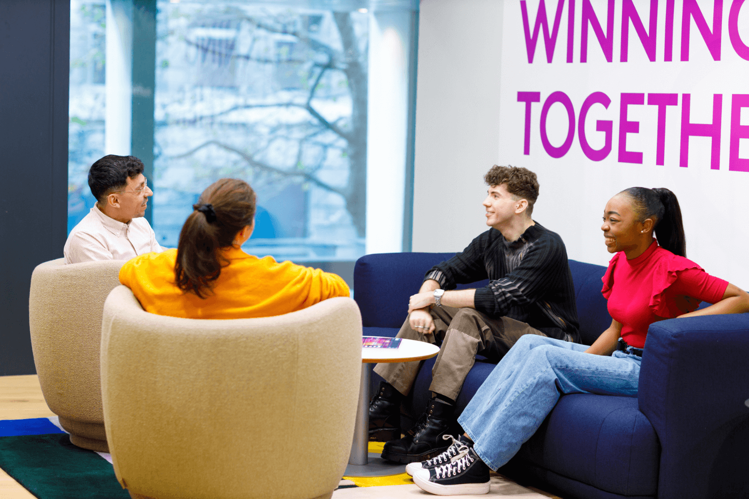 A group of four people sat on the sofa's all talking together; two men and two women. The wall behind them says "Winning Together"
